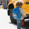 A little boy stand in front of a yellow school bus with a blue backpack.