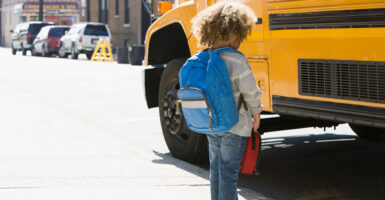 A little boy stand in front of a yellow school bus with a blue backpack.