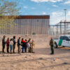 A group of about 10 illegal aliens stand near the border wall. Border Patrol agents wait with the group.