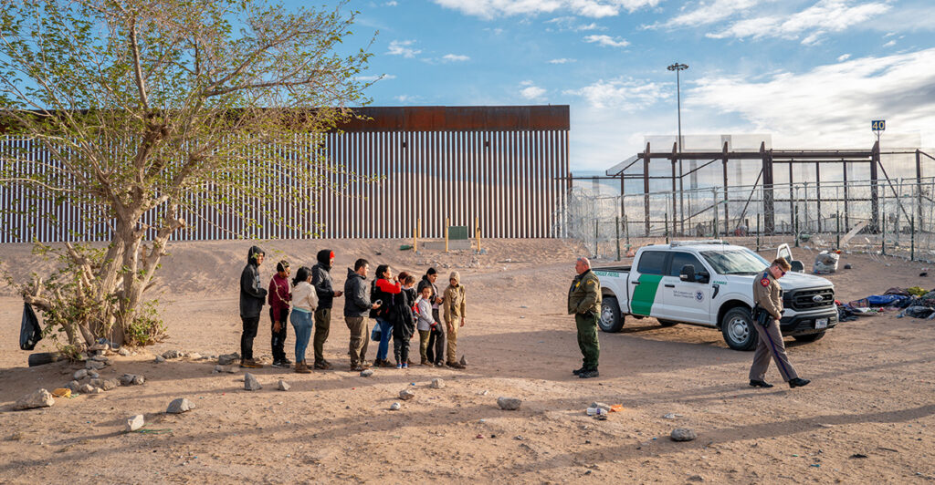 A group of about 10 illegal aliens stand near the border wall. Border Patrol agents wait with the group.