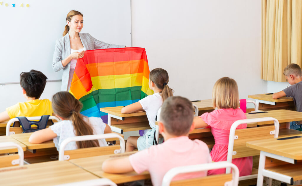 Young progressive female teacher discussing with preteen children about LGBT social movements in classroom, holding rainbow flag.