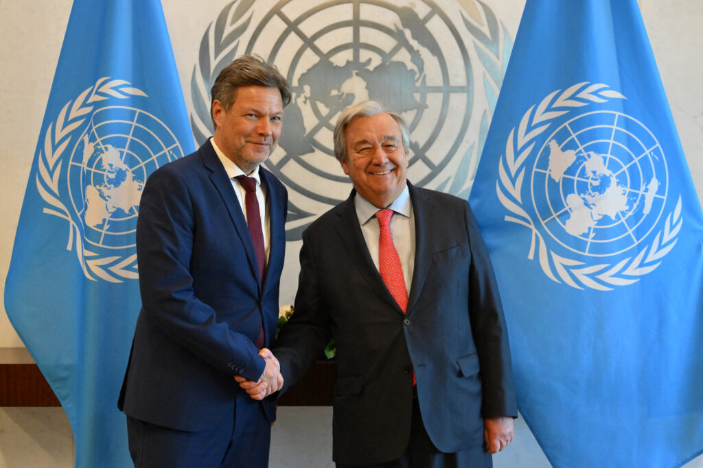Habeck and Gutteres wear suits and shake hands in front of United Nations flags.