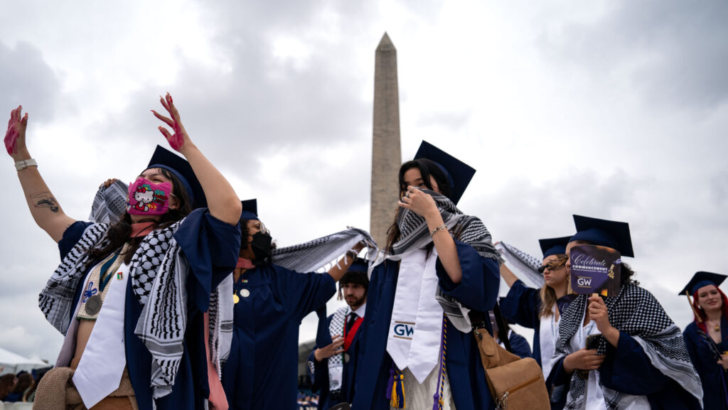 Students wear graduation caps and masks in front of the Washington Monument.