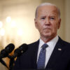 Joe Biden stands in front of a microphone wearing a suit and blue tie.
