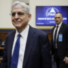 Attorney General Merrick Garland smiles as he enters a room, wearing a blue suit and tie.
