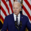 Joe Biden wears a navy-blue suit and speaks at a podium in front of American flags.