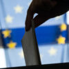 A woman's hand drops a paper ballot into a ballot box in front of the European Union flag.