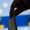 A woman's hand drops a paper ballot into a ballot box in front of the European Union flag.