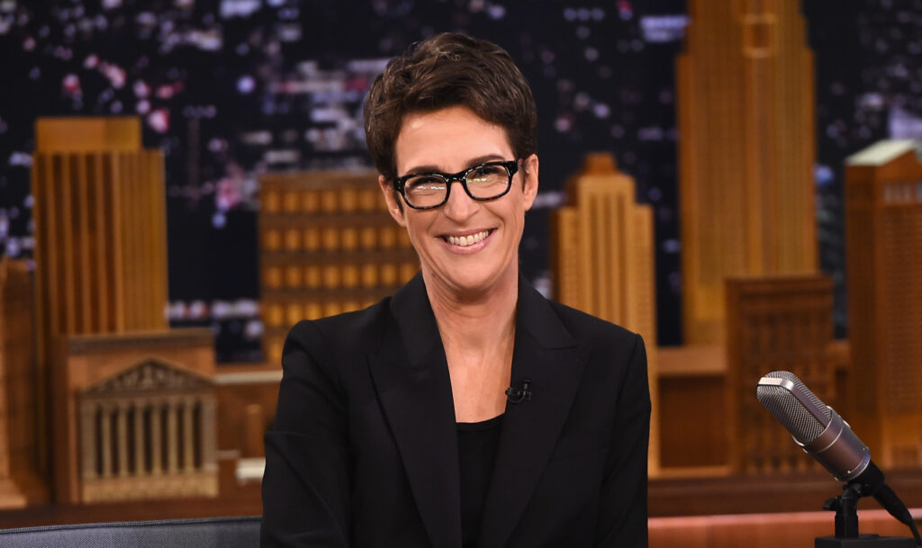 Rachel Maddow smiles at the camera and wears a black blazer and dark-rimmed glasses.