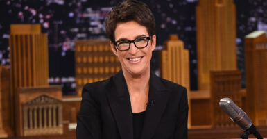 Rachel Maddow smiles at the camera and wears a black blazer and dark-rimmed glasses.