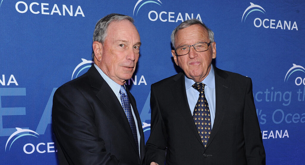 Swiss billionaire Hansjörg Wyss shakes hands with former New York Mayor Michael Bloomberg. Both wear suits in front of a blue banner.