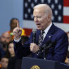 President Joe Biden in a blue suit raises a finger while speaking in a microphone