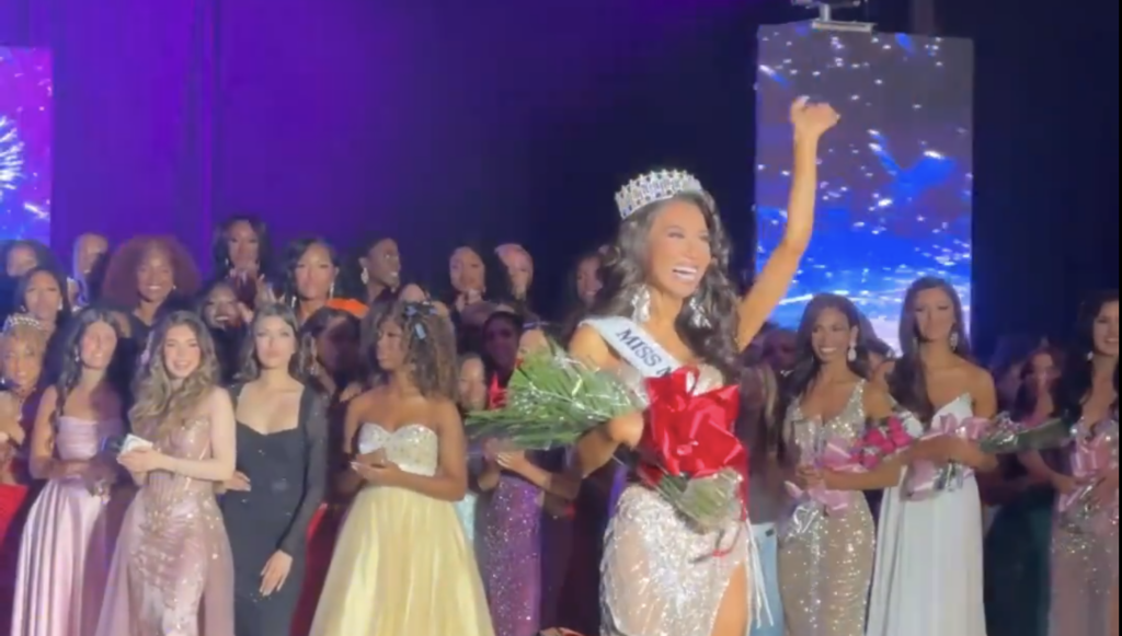 Trans-identifying male contestant waves waves from the stage in a gown, crown, and flowers with other contestants in the background