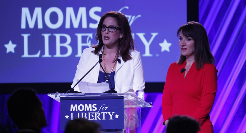 Tiffany Justice, in a blue dress and a white blazer, speaks in front of a podium reading "Moms for Liberty," as Tina Descovich, in a red dress, looks on.