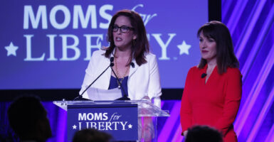 Tiffany Justice, in a blue dress and a white blazer, speaks in front of a podium reading "Moms for Liberty," as Tina Descovich, in a red dress, looks on.