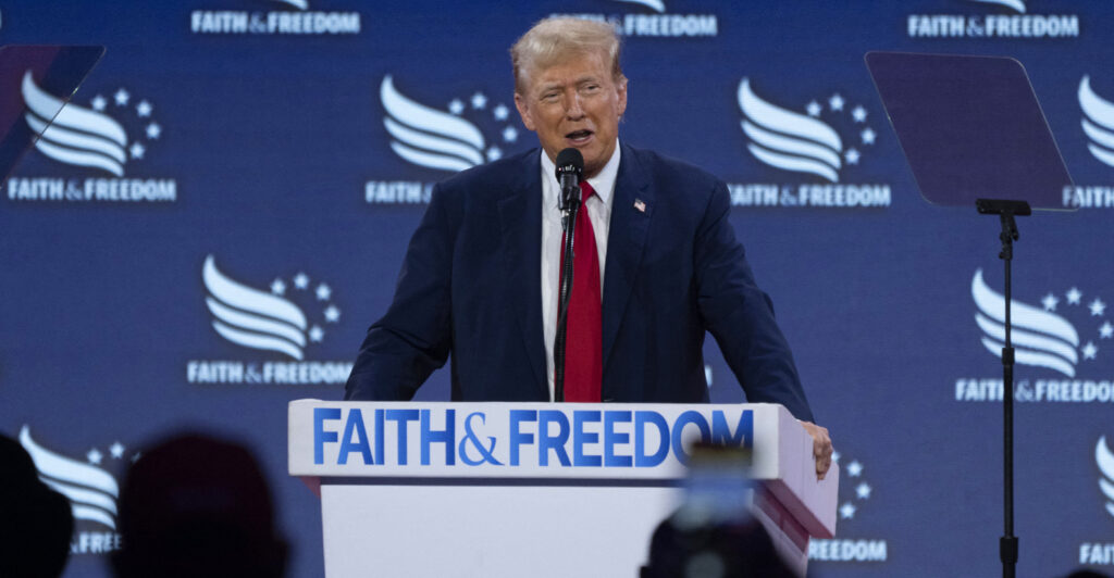 Donald Trump In a suit at a podium That says faith and freedom on the front in front of a blue background
