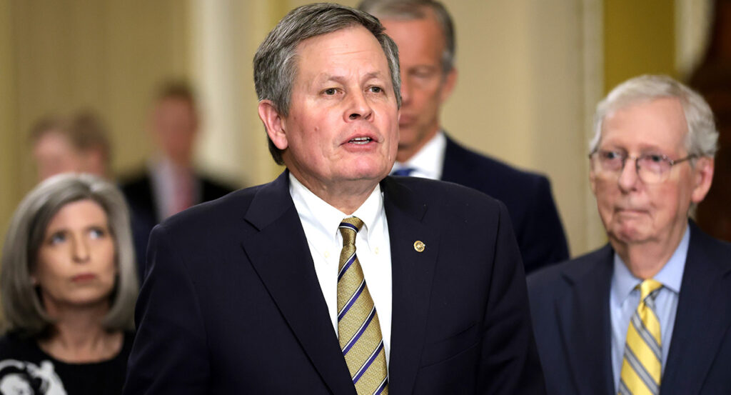 Sen. Steve Daines stands in front of several other senators in a suit and tie