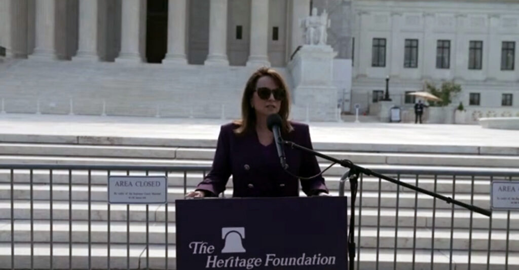 A woman speaks at a podium wearing sunglasses.