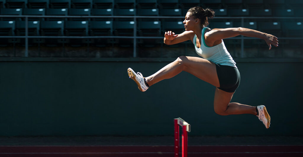 A female track athlete jumps over a hurdle.