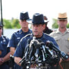 A sheriff speaks at a press conference with multiple microphones surrounded by other officers in uniform.