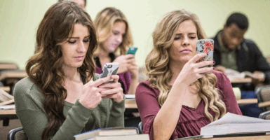 Two high school girls sitting in the classroom looking at their smart phones instead of paying attention