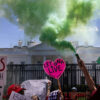 Protesters spraying green smoke and holding signs in front of the White House lawn.