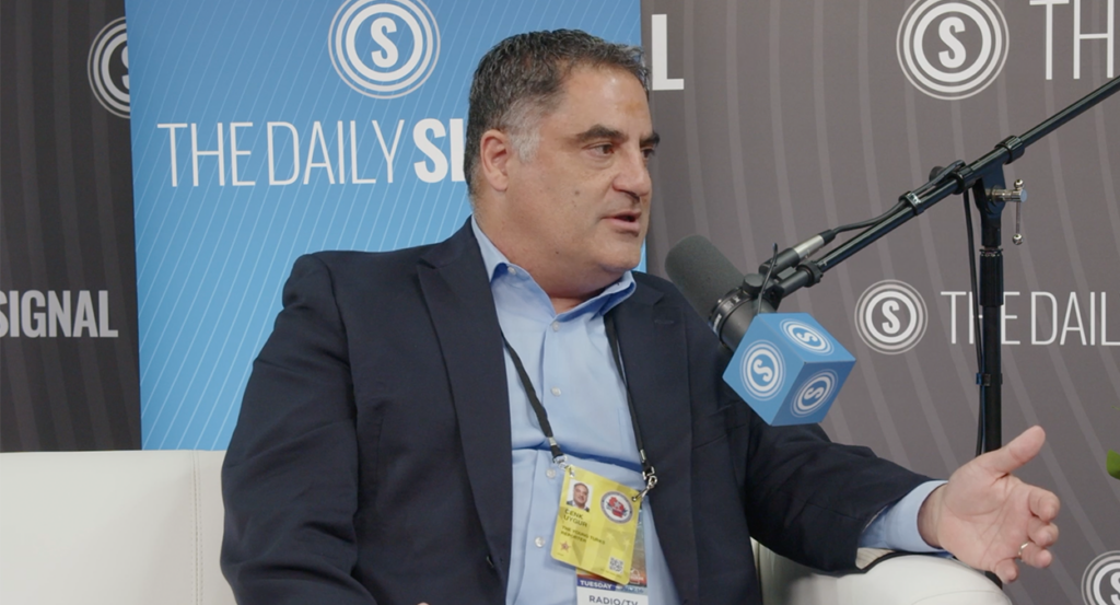 Cenk Uygur speaks at The Daily Signal booth wearing a suit without a tie