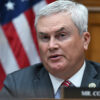 House Oversight Chairman James Comer in a suit sitting in a committee hearing in front of the American flag