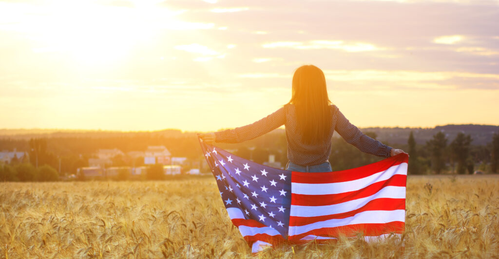 Silhouette of woman with the American flag in a wheat field against the sunset.