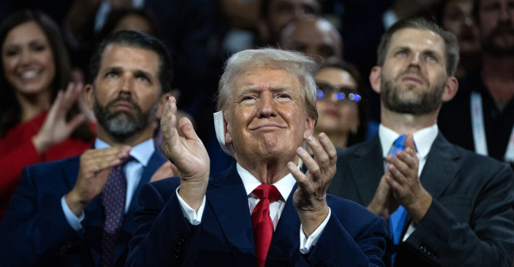 Former President Donald Trump, in a suit and clapping, pictured with his sons Don Jr. and Eric