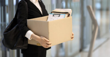 A woman wearing a pantsuit holds a box of her belongings.