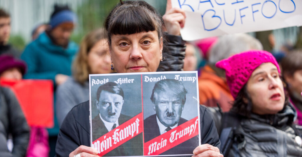 A woman holds a picture of Adolf Hitler and Donald Trump side-by-side, both with Hitler-style mustaches.
