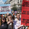 Protesters march with signs reading "Puberty Blockers Save Lives" and "Puberty Blockers for Cis Kids but Not Trans Kids"
