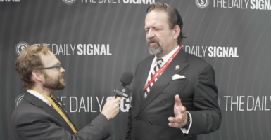 Tyler O'Neil in a grey suit holds the microphone for Seb Gorka in a black suit, gesturing.