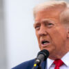 Donald Trump in a blue suit and a red tie at a microphone with an American flag in the background