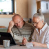 Senior couple sitting at the kitchen table with croissant and cups of tea, looking at digital tablet and recalculating their expenses.