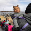 Migrant in black hoodie holds child in gray hoodie while crossing Rio Grande river with U.S. border wall in background.