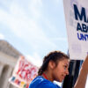 A pro-life supporter stands on a lamppost and holds a sign that reads “MAKE ABORTION UNTHINKABLE” in front of the Supreme Court.