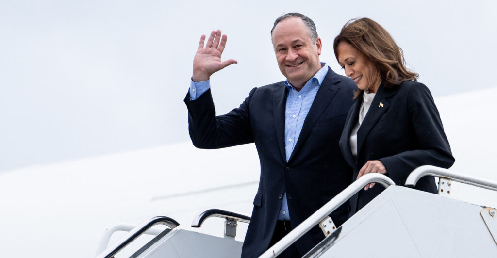 Second Gentleman Doug Emhoff waves in a suit without a tie as Kamala Harris descends stairs