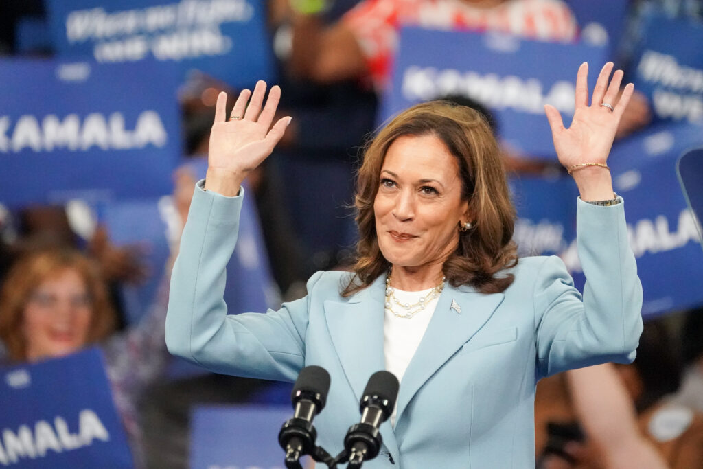 Kamala Harris wears a blue blazer and hold both hands up to a crowd.