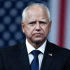 Walz stares grimly at the camera with a U.S. flag hanging behind him.