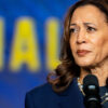 Kamala Harris Behind microphones in front of a blue background with yellow lettering