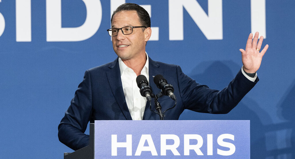 Josh Shapiro in a blue suit raises his hind behind a podium reading "Harris"