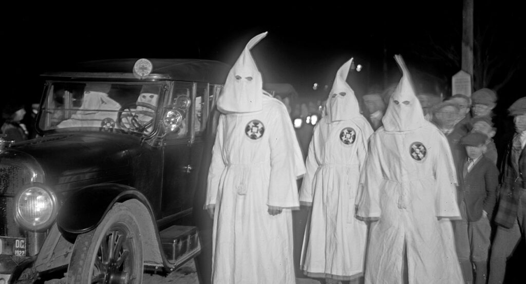 Ku Klux Klan members in white robes marching near an old-fashioned vehicle