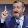 Ted Cruz, in a blue suit with a blue tie, raises his finger in front of a picture of the U.S. Capitol