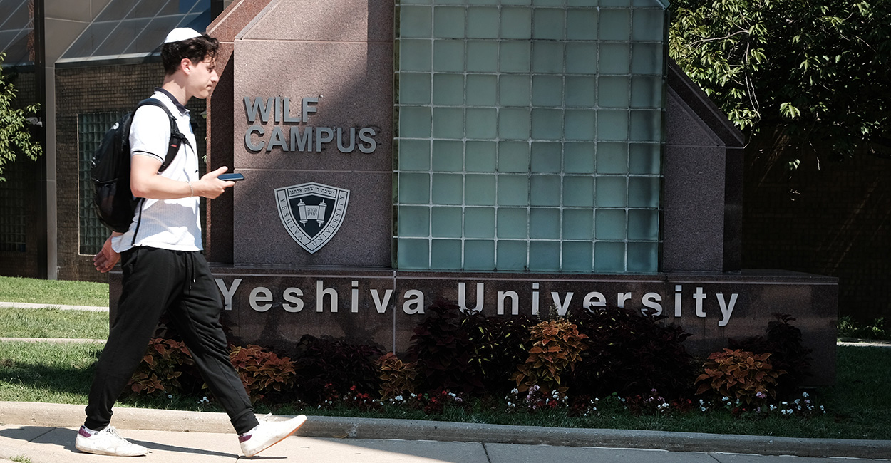 Student wearing a backpack walks in front of a Yeshiva University sign